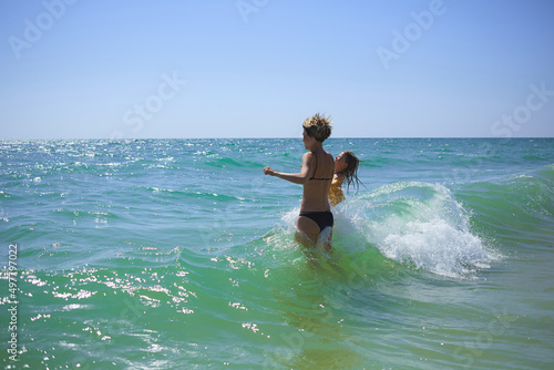 Summer happy family of six years blonde child playing and jumping water waves embracing woman mother in sea shore beach © sarymsakov.com