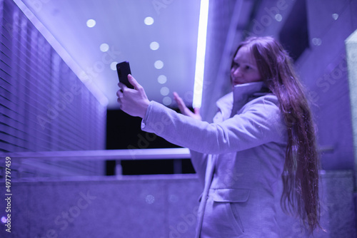 Young millennial woman using mobile phone video chatting on social media or doing work video call standing near office modern architecture building with purple blue violet neon light at night
