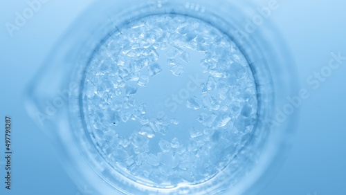 Top view macro shot of crystals dissolving in the beaker with transparent liquid on pale blue background | Mineral skincare cosmetics formulating concept