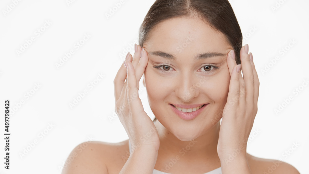 Pretty Caucasian young woman massages her temples stretching skin around eyes and smiles on white background | Droopy eyes removal concept