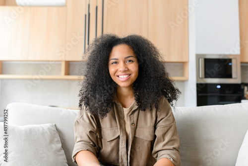 Portrait of serene and charming beautiful dark-haired woman. Pretty multiracial lady in casual wear looks at the camera and smiles, sitting on the comfortable white sofa