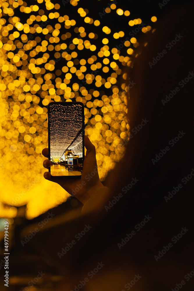 Closeup of a woman at night taking a picture on mobile phone camera of golden yellow lights in the street in city, real people using technology and mobile photography hobby Closeup of a woman at night taking a picture on mobile phone camera of golden yellow lights in the street in city, real people using technology and mobile photography hobby