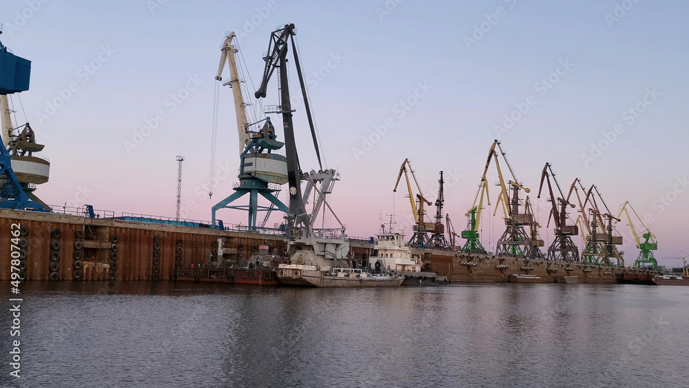 The large cranes at the sea port on sunset sky background. Clip. Big industrial machines and calm water surface, concept of goods transportations.