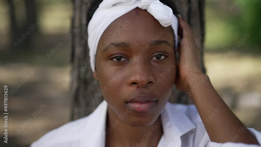 Close-up front view of confident young African American woman looking at camera leaning on tree trunk in forest. Portrait of relaxed carefree lady posing in slow motion travelling outdoors Close-up front view of confident young African American woman looking at camera leaning on tree trunk in forest. Portrait of relaxed carefree lady posing in slow motion travelling outdoors