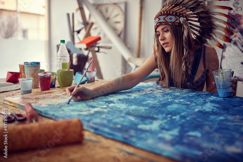 Valokuva A young sexy female artist with a war bonnet is in ecstasy while working on a new painting in her studio