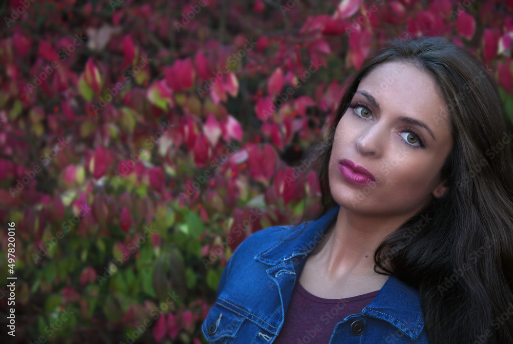 young woman in her twenties with long natural black hair posing outside in front of red flowers with blue jeans jacket