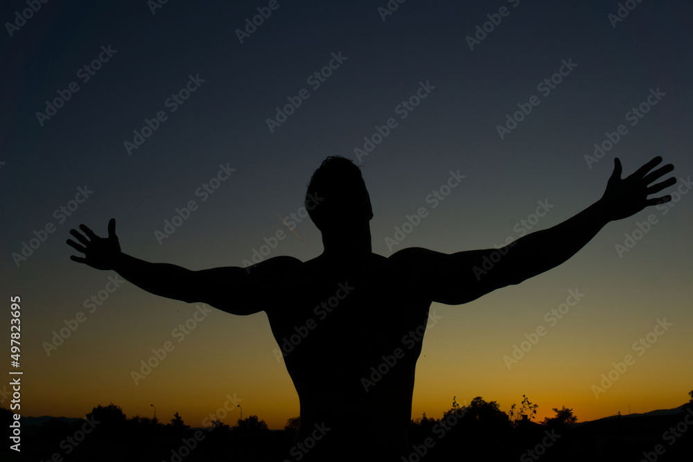 Silhouette of muscular man posing shows his muscles against the sky at sunset. 