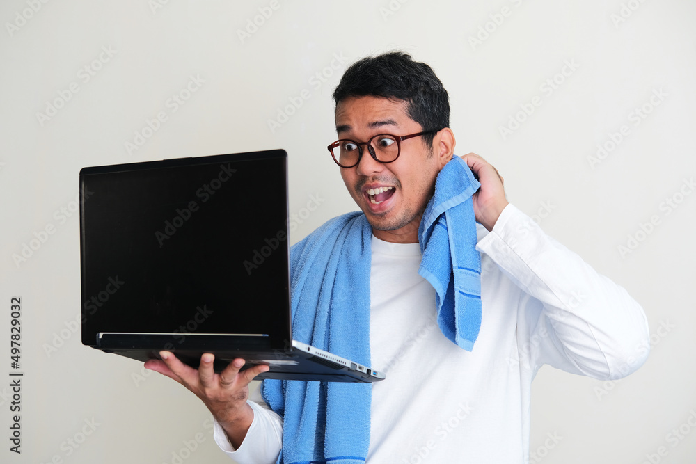 Adult Asian man doing online meeting using laptop while still wearing sleeping clothes and towel around his neck Adult Asian man doing online meeting using laptop while still wearing sleeping clothes and towel around his neck