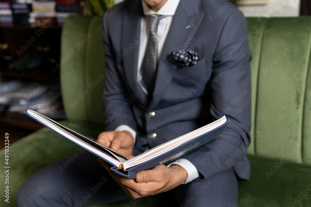 Man in expensive tailored suit sitting on sofa, choosing material from catalogue in studio. Man in expensive tailored suit sitting on sofa, choosing material from catalogue in studio.
