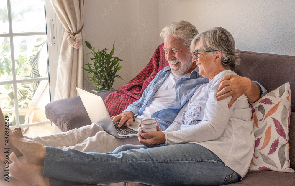 Cheerful senior couple at home on sofa using laptop computer. Two elderly retirees enjoying free time and technological devices Cheerful senior couple at home on sofa using laptop computer. Two elderly retirees enjoying free time and technological devices