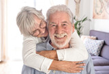 Beautiful senior caucasian woman lovingly hugging her seated husband. Smiling elderly couple white haired looking at camera