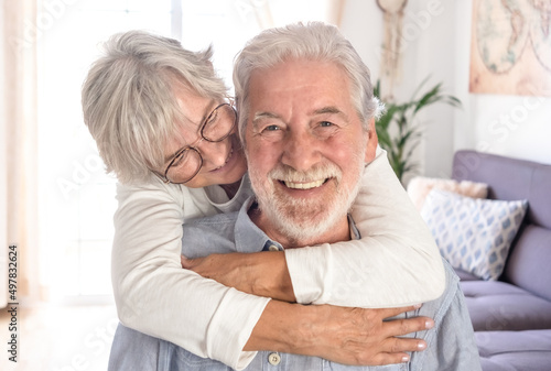 Beautiful senior caucasian woman lovingly hugging her seated husband. Smiling elderly couple white haired looking at camera