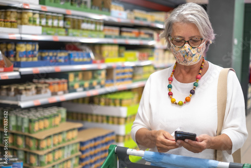 Attractive elderly woman makes purchases in the supermarket by pushing the cart and looking at mobile phone, wearing a protective mask