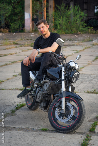 A young motorcyclist in a black T-shirt sits on a classic naked motorcycle parked in an abandoned area