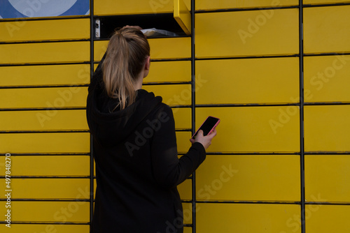 Beautiful woman picking up a package from a smart electronic steel parcel locker box, automatic mailboxes. Paczkomat delivery service, collection machine. Using app on her phone to open the door. photo