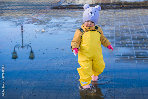 A child aged 2-3 years old dressed in a yellow rubber jumpsuit walks through puddles in the park