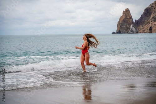 A beautiful and sexy brunette in a red swimsuit on a pebble beach, Running along the shore in the foam of the waves