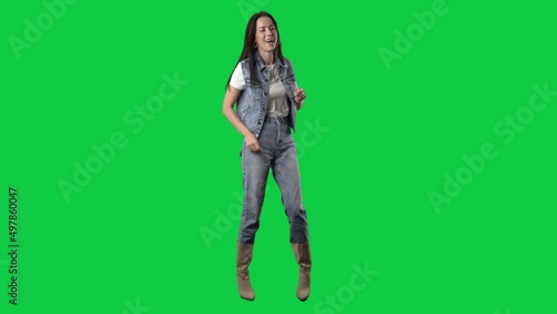 Cheerful young woman in jeans and boots singing and dancing carefree. Full body isolated on green screen background