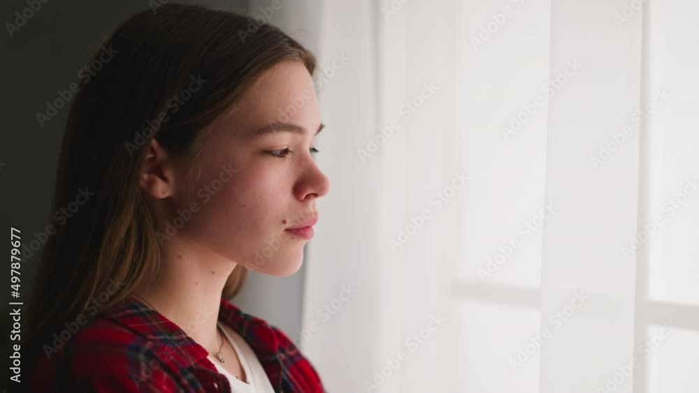 Depressed young woman with long brown hair stands looking out of window in light room closeup slow motion. Psychotherapy to help change mental state Depressed young woman with long brown hair stands looking out of window in light room closeup slow motion. Psychotherapy to help change mental state