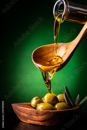 from the wooden spoon, in the foreground, the extra virgin olive oil is poured into an olive wood bowl with olives