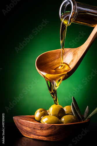 from the wooden spoon, in the foreground, the extra virgin olive oil is poured into an olive wood bowl with olives photo