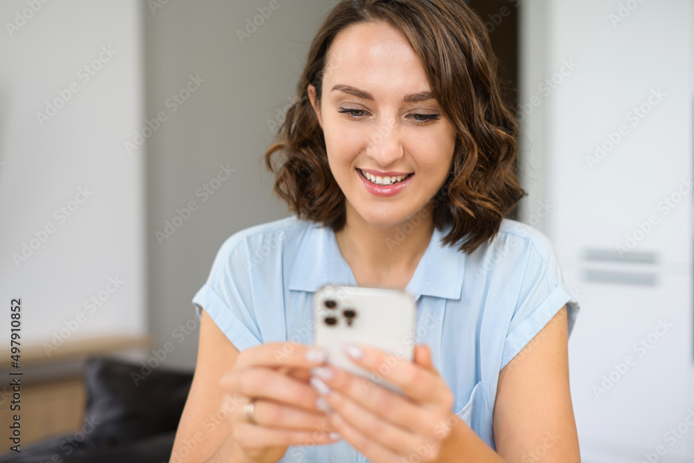 Portrait view of the calm young woman in casual wear sitting on the bed in cozy bedroom and holding smartphone. Female spending time online, texting with friends Portrait view of the calm young woman in casual wear sitting on the bed in cozy bedroom and holding smartphone. Female spending time online, texting with friends