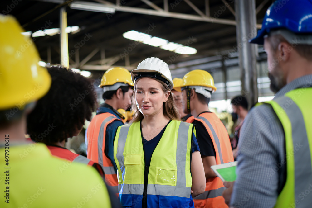 Obraz premium A confident, beautiful Caucasian woman worker stands among the male workers wearing safety uniform are meeting morning talk before the start of work in heavy industrial manufacturer factory.