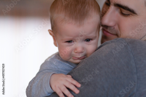 Closeup Sad young blond boy crying on father hands in room. Man holds son, hugs and comforts. Family love, care and moral support, baby's tears, daddy's arms. Dad consoling crying child