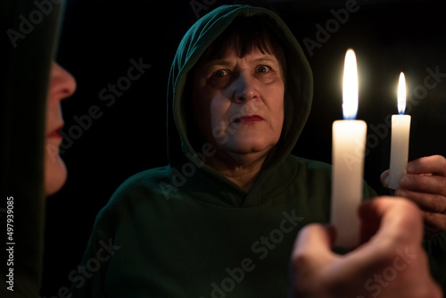 An elderly woman with a candle in her hands looks at her reflection in the mirror.