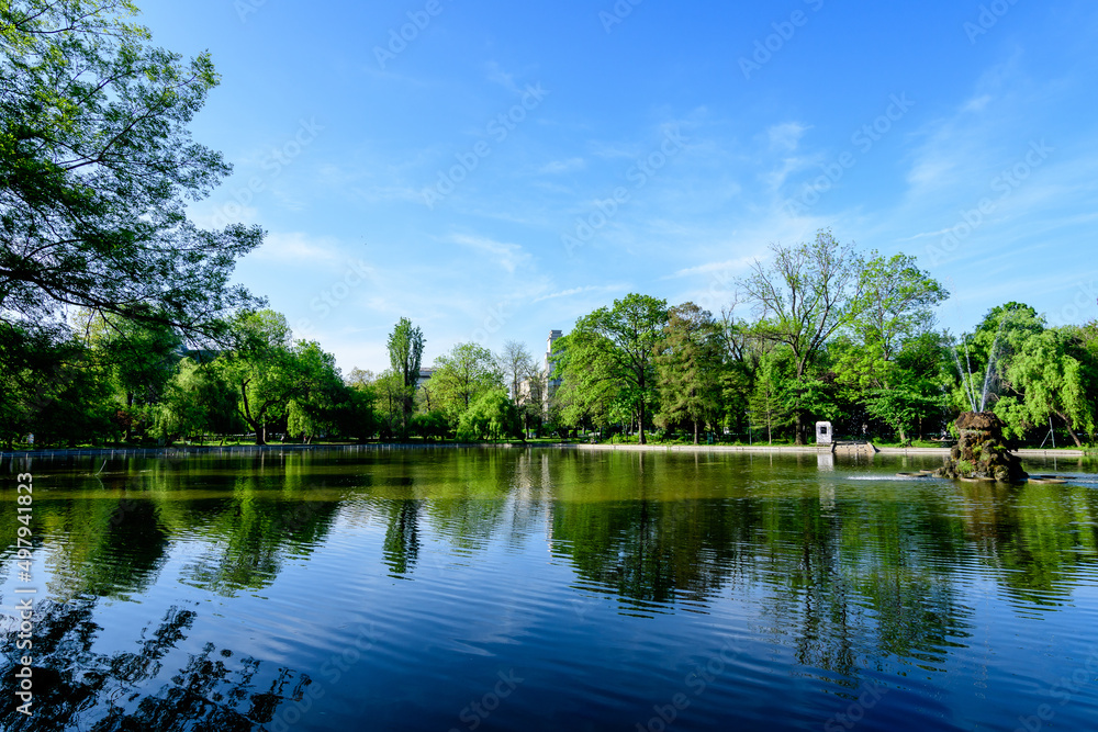 Vivid green landscape with old large linden trees and small boats near the lake in Cismigiu Garden (Gradina Cismigiu), a public park in the city center of Bucharest, Romania, in a sunny spring day.