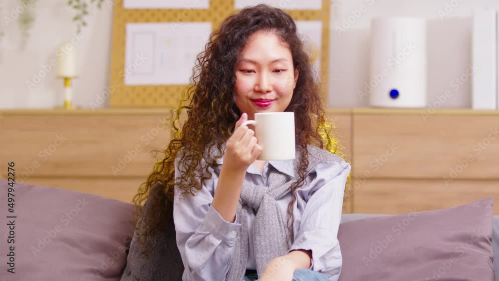 Asian peaceful woman holding cup enjoy favorite tea or coffee morning drink while sitting at sofa in modern living room before start work. Break, pause, daydreaming and work life balance. Asian peaceful woman holding cup enjoy favorite tea or coffee morning drink while sitting at sofa in modern living room before start work. Break, pause, daydreaming and work life balance.