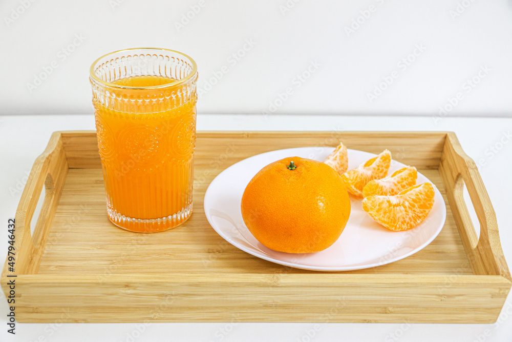 Naklejka premium Glass of sweet and natural orange juice, full of vitamin goodness, besides full round orange and peeled orange pieces in a wooden tray against white background