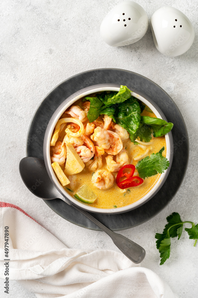 Asian laksa soup with shrimp in a ceramic bowl on a light culinary background. Spicy broth soup with coconut milk, rice noodles and seafood in a plate on the closeup kitchen table. Top view