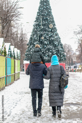 Young happy cheerful family near the Christmas tree in winter afternoon. Boy sits on the father shoulders. Woman hugs husband  and child.  rear view of walking people