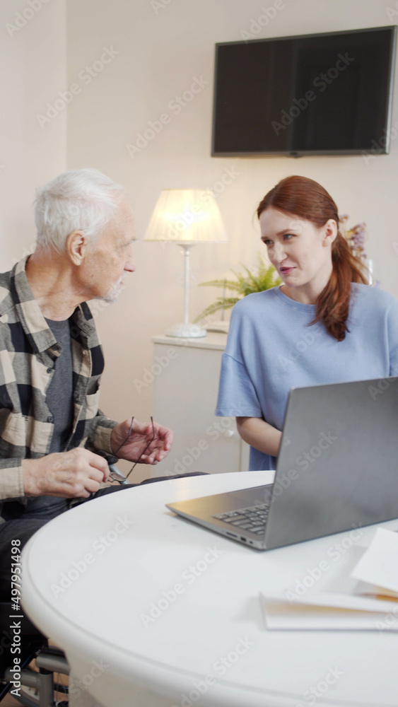An elderly man in a wheelchair and a young woman sit at a table and chat An elderly man in a wheelchair and a young woman sit at a table and chat