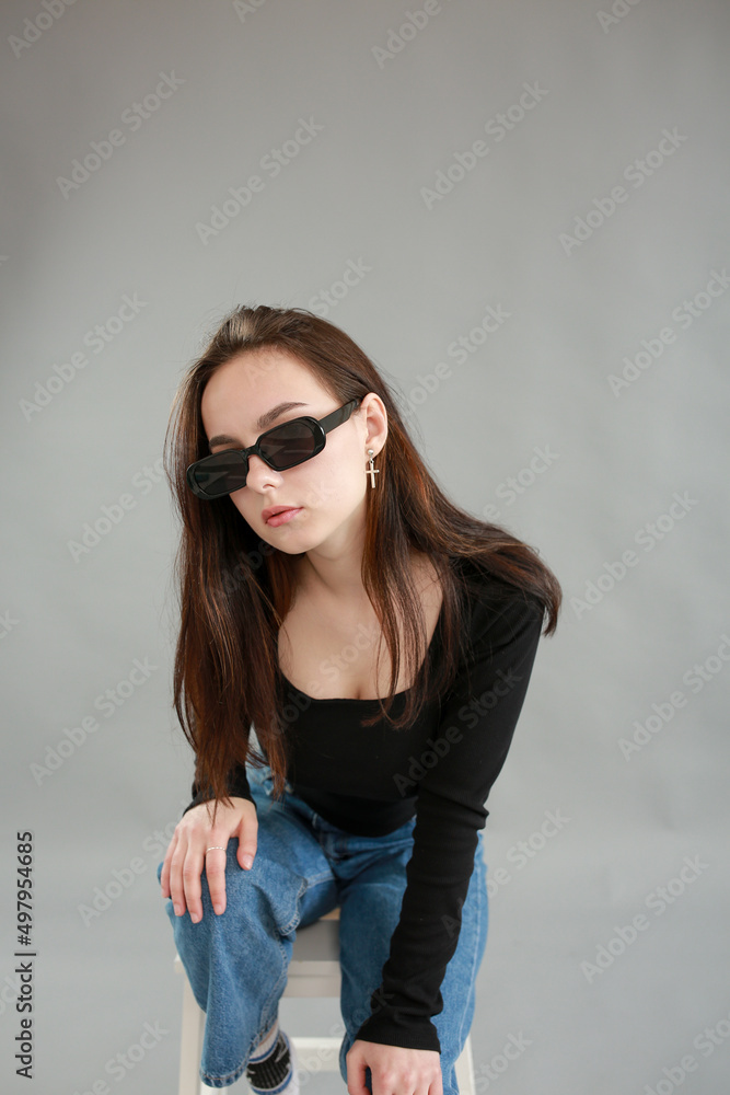 beautiful young woman on a gray background in black sunglasses. sit on a white chair. poses for a photo shoot beautiful young woman on a gray background in black sunglasses. sit on a white chair. poses for a photo shoot