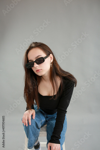 beautiful young woman on a gray background in black sunglasses. sit on a white chair. poses for a photo shoot © Anhelina Tyshkovets