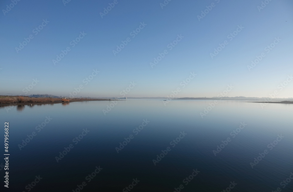 A tranquil early morning view across a still lake under a clear blue sky. A tranquil early morning view across a still lake under a clear blue sky.