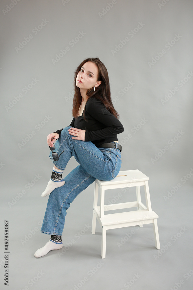 beautiful young woman on a gray background in black sunglasses. sit on a white chair. poses for a photo shoot beautiful young woman on a gray background in black sunglasses. sit on a white chair. poses for a photo shoot
