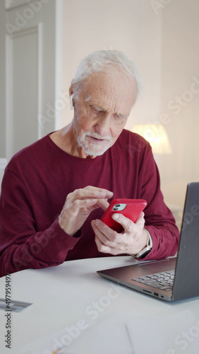 An elderly man sits at a table in the living room and flips through photos on a smartphone