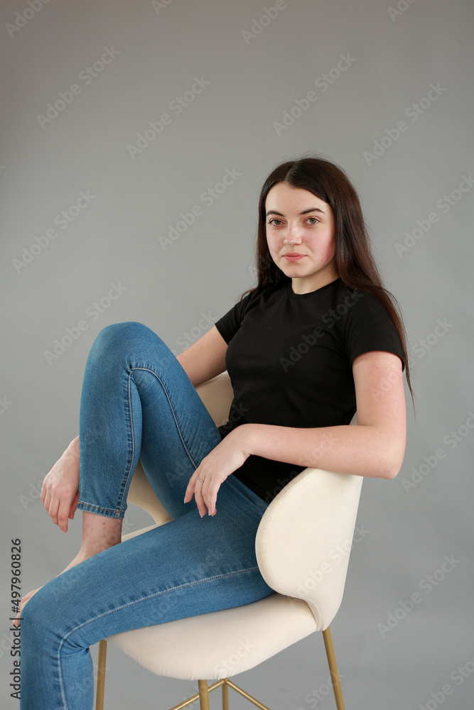 stylish photo shoot beautiful model with black hair. girl in jeans and black turtleneck on a gray background. girl sitting on a white chair and posing for a photo stylish photo shoot beautiful model with black hair. girl in jeans and black turtleneck on a gray background. girl sitting on a white chair and posing for a photo