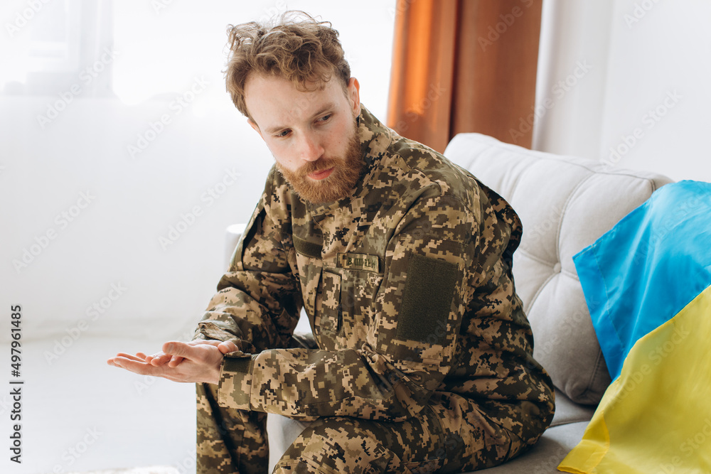Portrait of an emotional young Ukrainian patriot soldier in military uniform sitting on the office on a sofa with a yellow and blue flag.