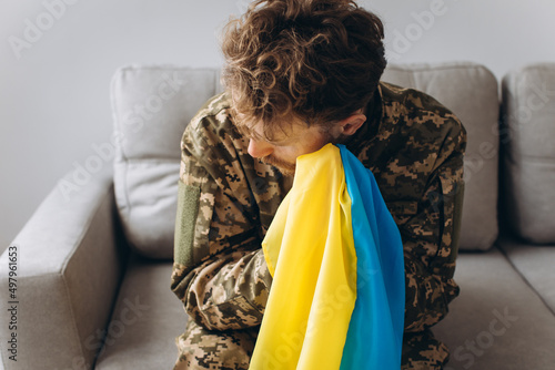 Portrait of an emotional young Ukrainian patriot soldier in military uniform sitting on the office on the couch holding a yellow and blue flag. photo