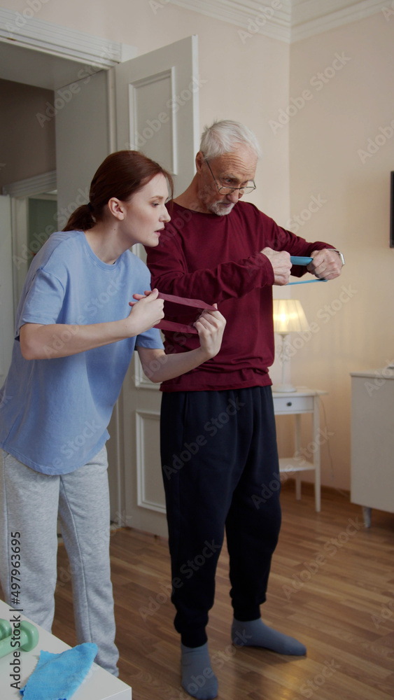 Fototapeta premium A woman puts a fitness band in the hands of an elderly man and tells how to use it during a workout