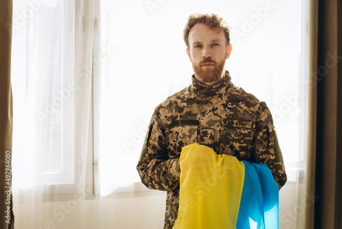 Portrait of an emotional young bearded Ukrainian patriot soldier in military uniform holding a flag in the office