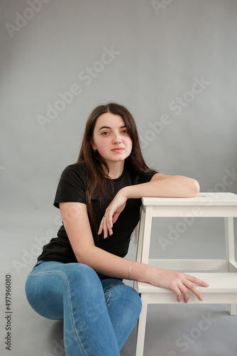 stylish photo shoot beautiful model with black hair. girl in jeans and black turtleneck on a gray background. girl sitting on a white chair and posing for a photo