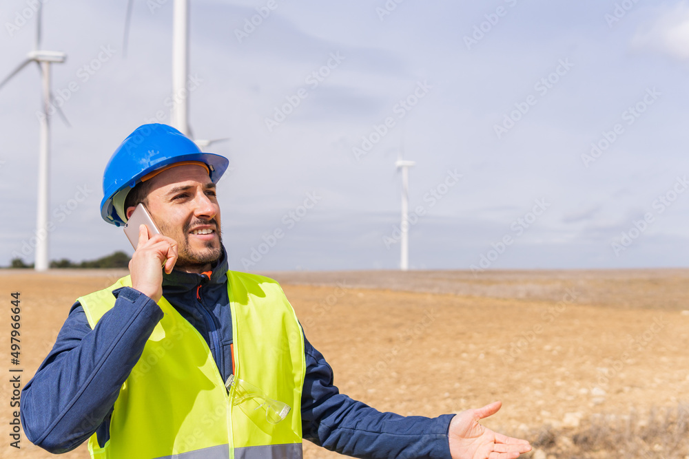 © Dani D.G - Adult man dressed in work clothes, safety helmet and reflective vest, smiling, talking on his mobile phone with his co-worker at a wind farm. Security technician in a field with wind generators.