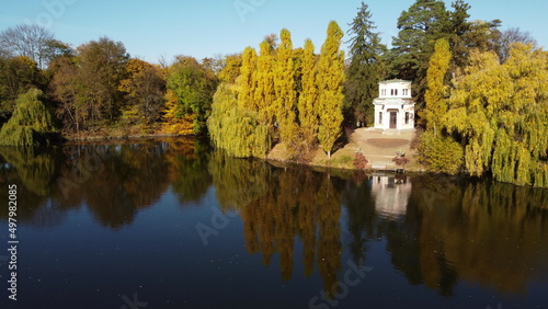 Aerial Drone View Flight Over water mirror surface of lake in park on windless sunny autumn day. Trees with yellow leaves on banks. In middle of lake - white motor boat. White building on the coast