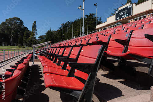 Long view looking down a row of red and black seats in an outdoor stadium with part of the track visible below and the scoreboard above