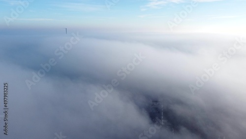 Aerial drone view flight over fog covering city. Scenic aerial view moving white foggy clouds and blue sky. Fog fast moving above rooftops of city houses. Sky landscape. Nature background. Environment